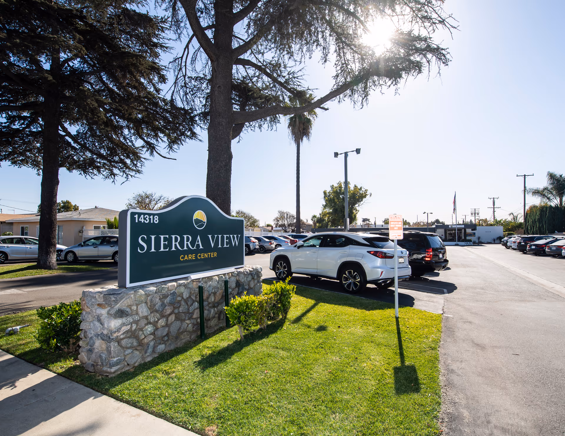 Outdoor view of Sierra View Care Center showing a green sign with the facility name and address 14318, a parking lot with several cars, trees, and a clear sky with sunlight.