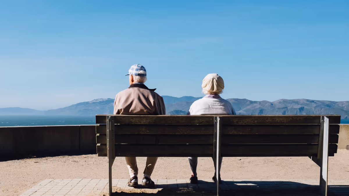 Two elderly people sit on a wooden bench overlooking the sea and mountains.
