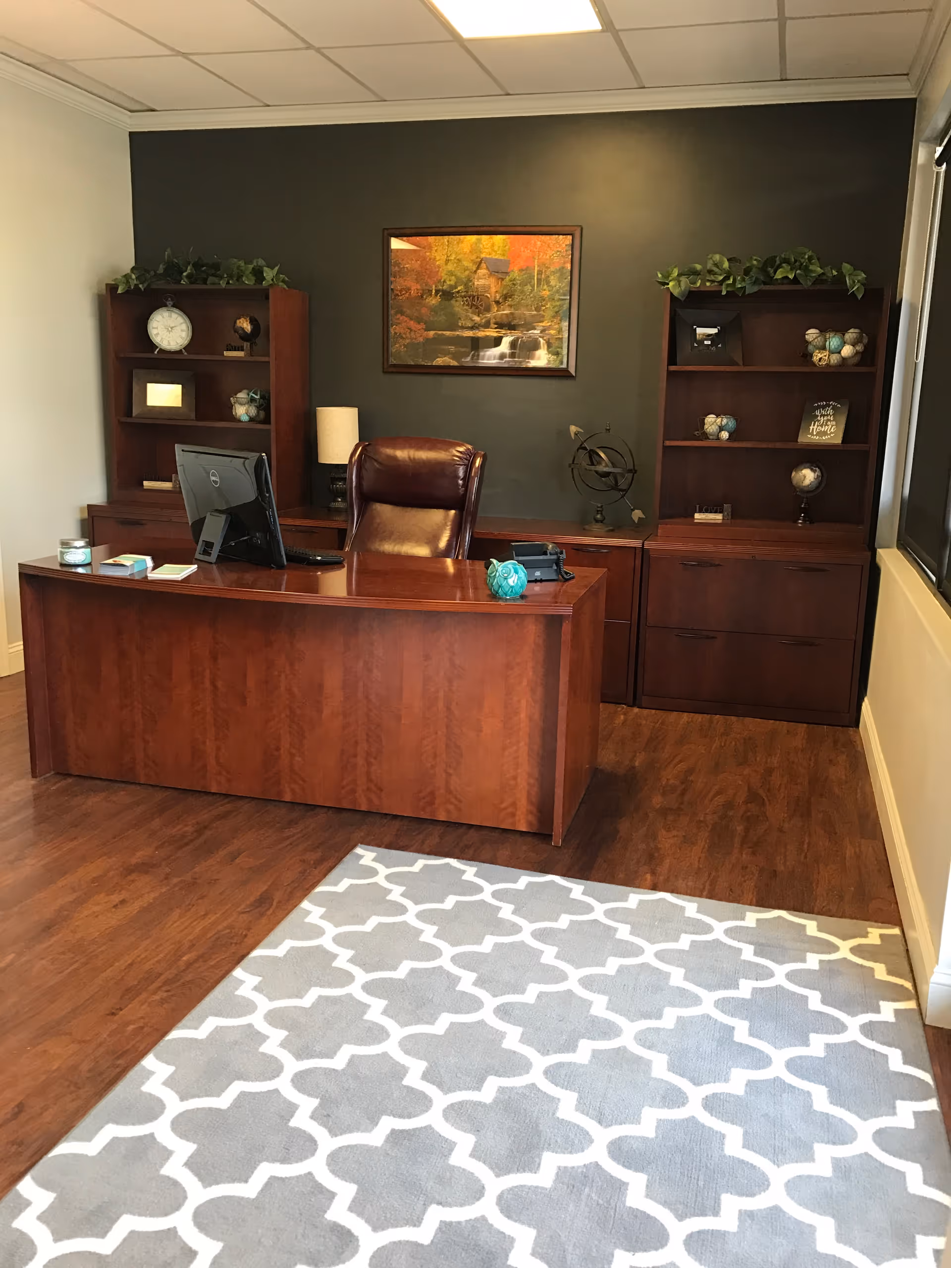 A wood-toned executive desk with computer, surrounded by bookcases, wall art, and a patterned rug.
