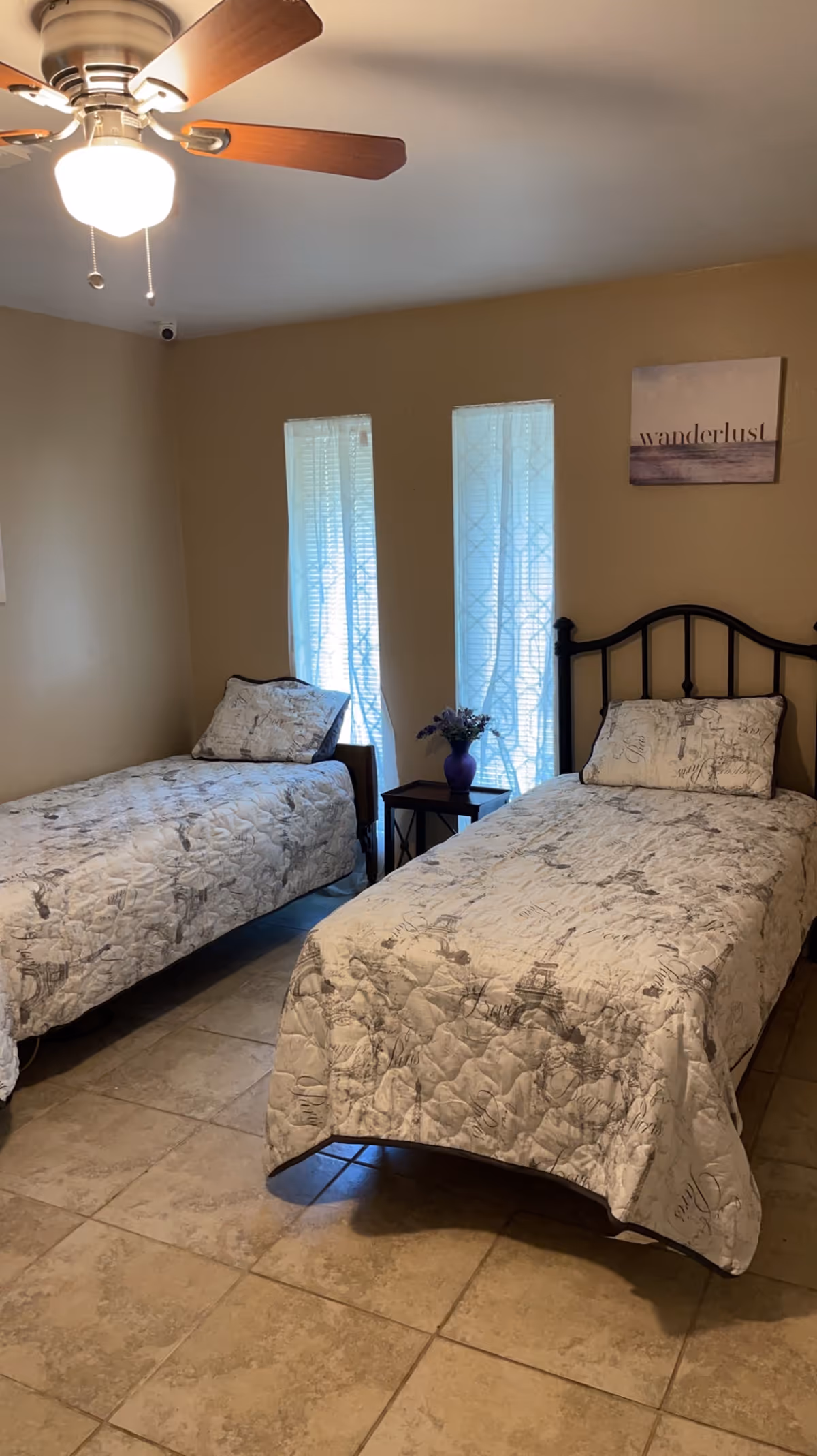 A bedroom with two twin beds covered in matching light-colored quilts with a subtle pattern. Between the beds is a small dark wooden nightstand with a purple vase holding flowers. Two narrow windows with sheer curtains allow natural light into the room. A ceiling fan with a light fixture is visible above. On the wall above one bed is a framed picture with the word 'wanderlust'.