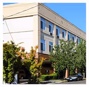 Two-story beige building with blue-trimmed windows, parked cars, and trees along the front.