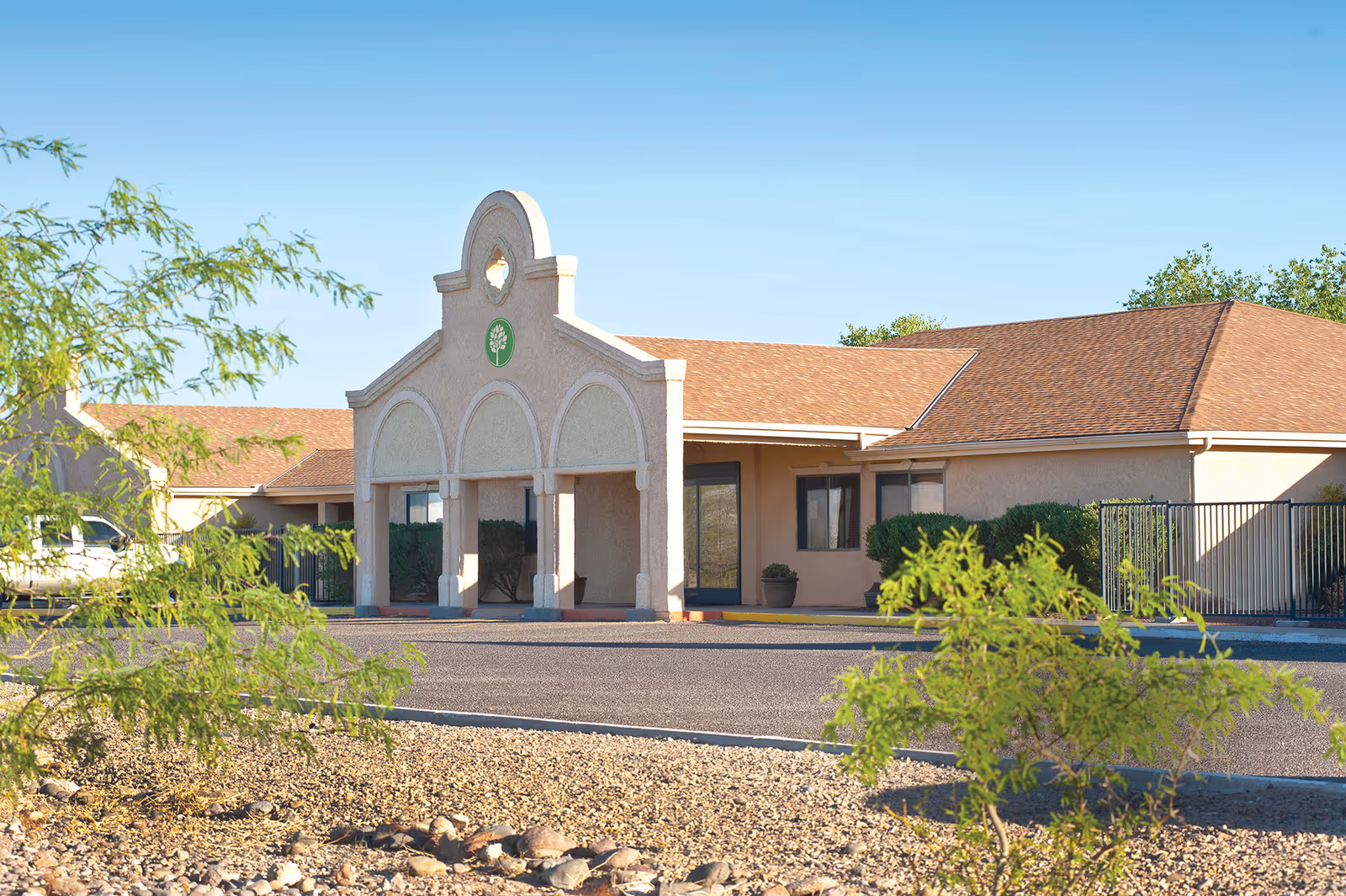 Exterior view of a single-story building with a beige facade and a brown shingle roof under a clear blue sky. The building has an arched entrance with columns and a circular emblem featuring a tree. There are some green bushes and small trees in the foreground along with a gravel area and a paved driveway.