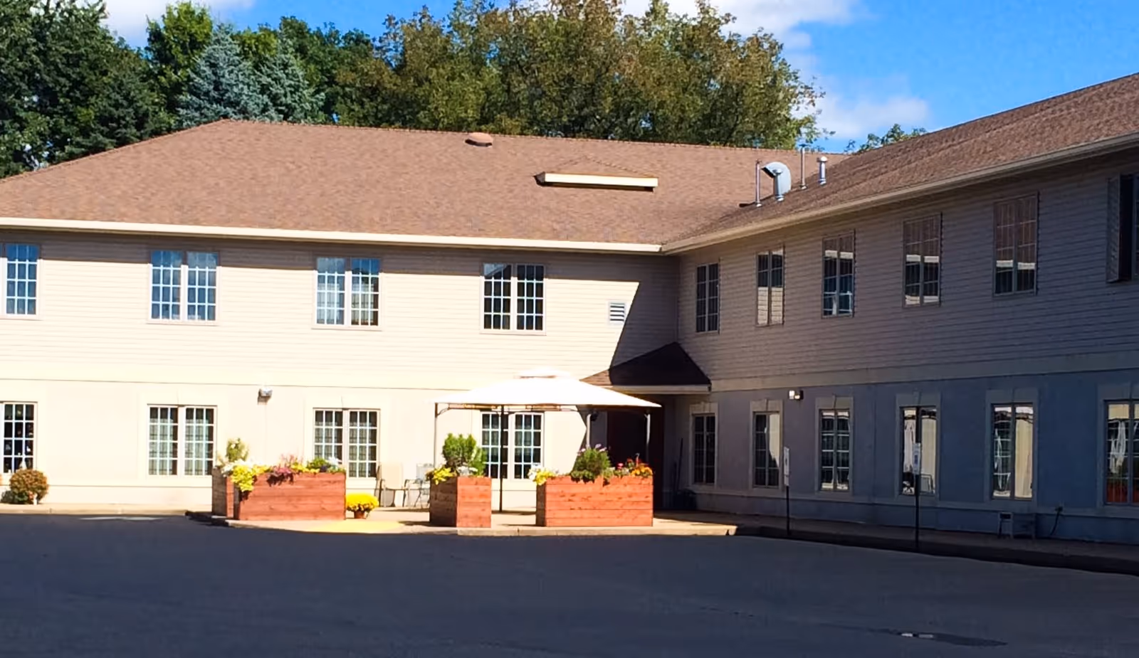 Exterior view of a two-story senior living facility building with beige siding and multiple windows. In front of the building, there are large wooden planters with flowers and a white umbrella providing shade over a seating area. Trees and a blue sky with some clouds are visible in the background.