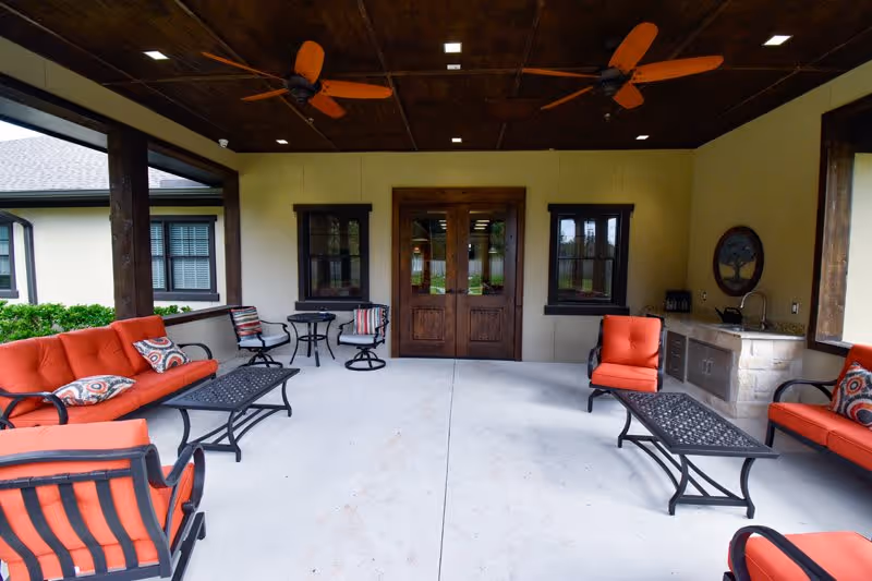 Covered outdoor patio area with orange cushioned seating including sofas and chairs, two ceiling fans, a small table with two chairs, and a built-in sink and counter on the right side. The patio has a concrete floor and wooden ceiling with recessed lighting. Double wooden doors and two windows are visible on the far wall.