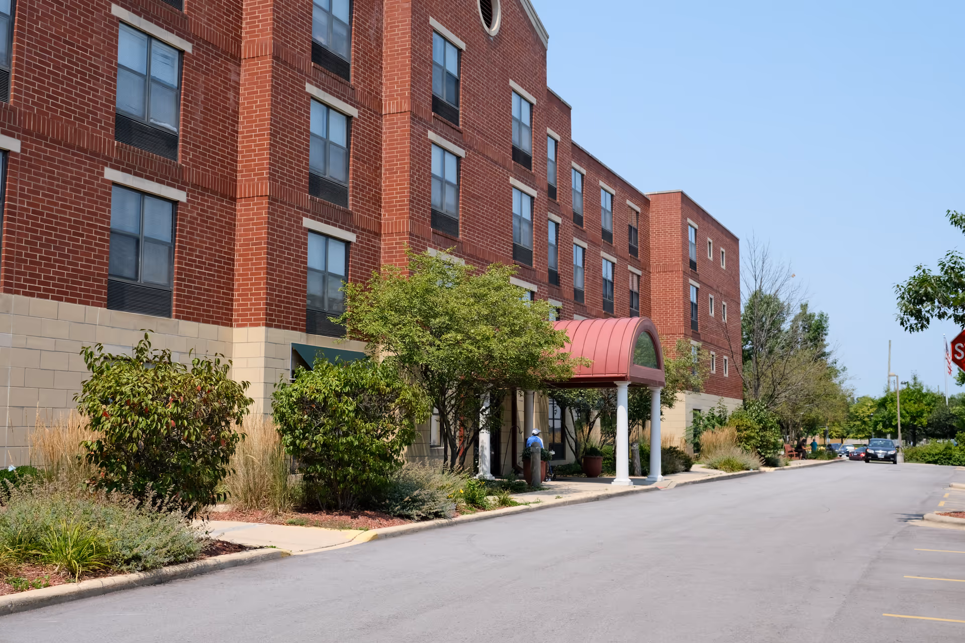 Exterior view of a multi-story brick building with a beige lower facade and multiple windows. There is a covered entrance with a red arched roof supported by white columns. Shrubs and trees line the sidewalk next to the building, and a few cars are parked along the street under a clear blue sky.