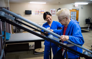 A healthcare worker in blue scrubs assists an elderly woman wearing a blue jacket as she uses parallel bars for physical therapy in a rehabilitation room.