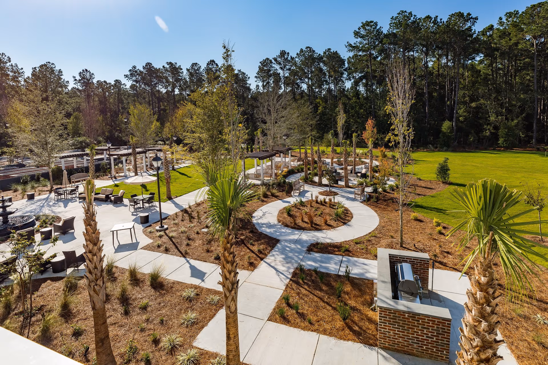 Outdoor garden area at Merrill Gardens at Carolina Park featuring paved walkways, palm trees, seating areas with tables and chairs, pergolas, a brick barbecue grill, and a backdrop of tall trees under a clear sky.