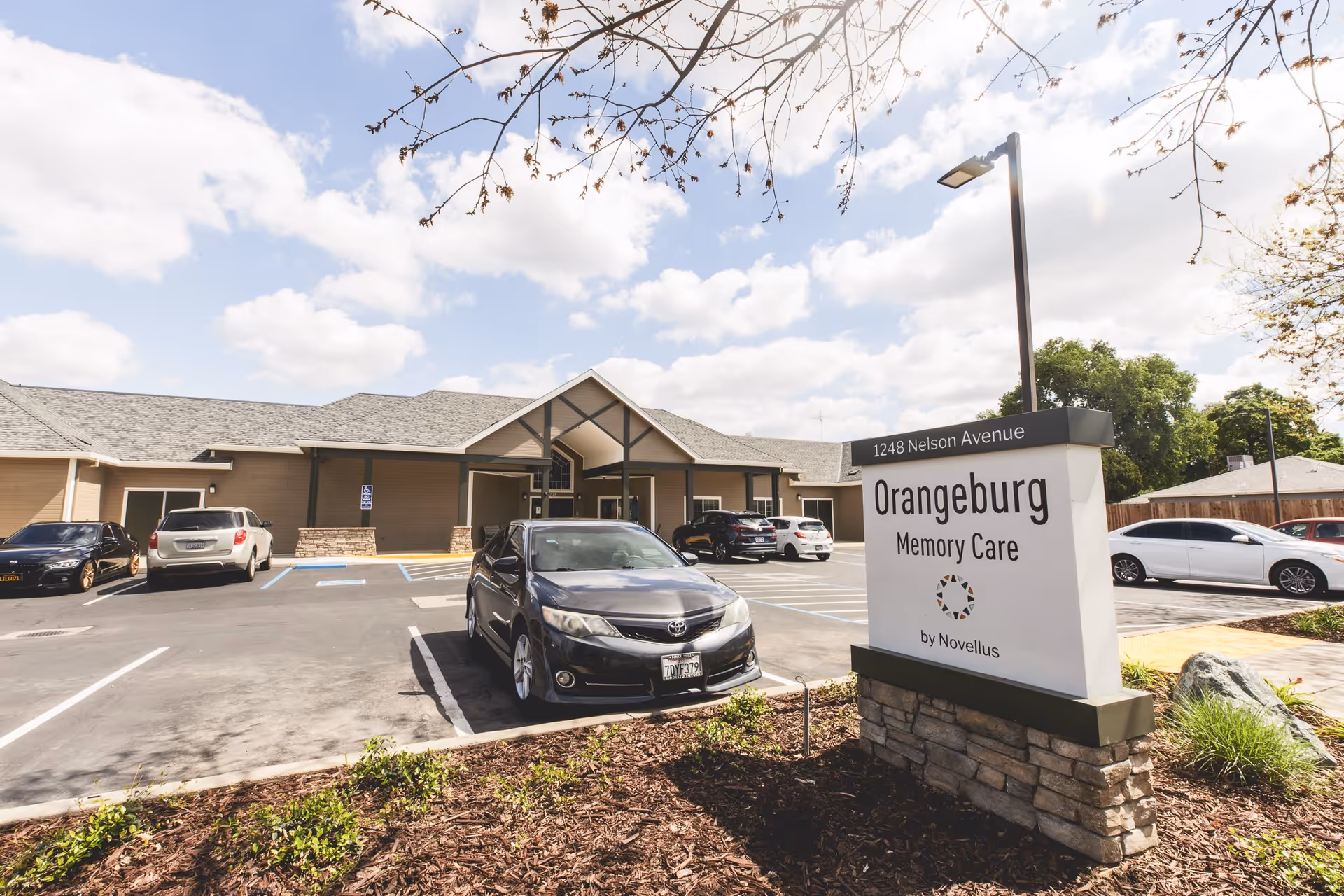 Exterior view of Orangeburg Memory Care facility showing the building entrance, parking lot with several cars, and a sign displaying the facility name and address at 1248 Nelson Avenue.