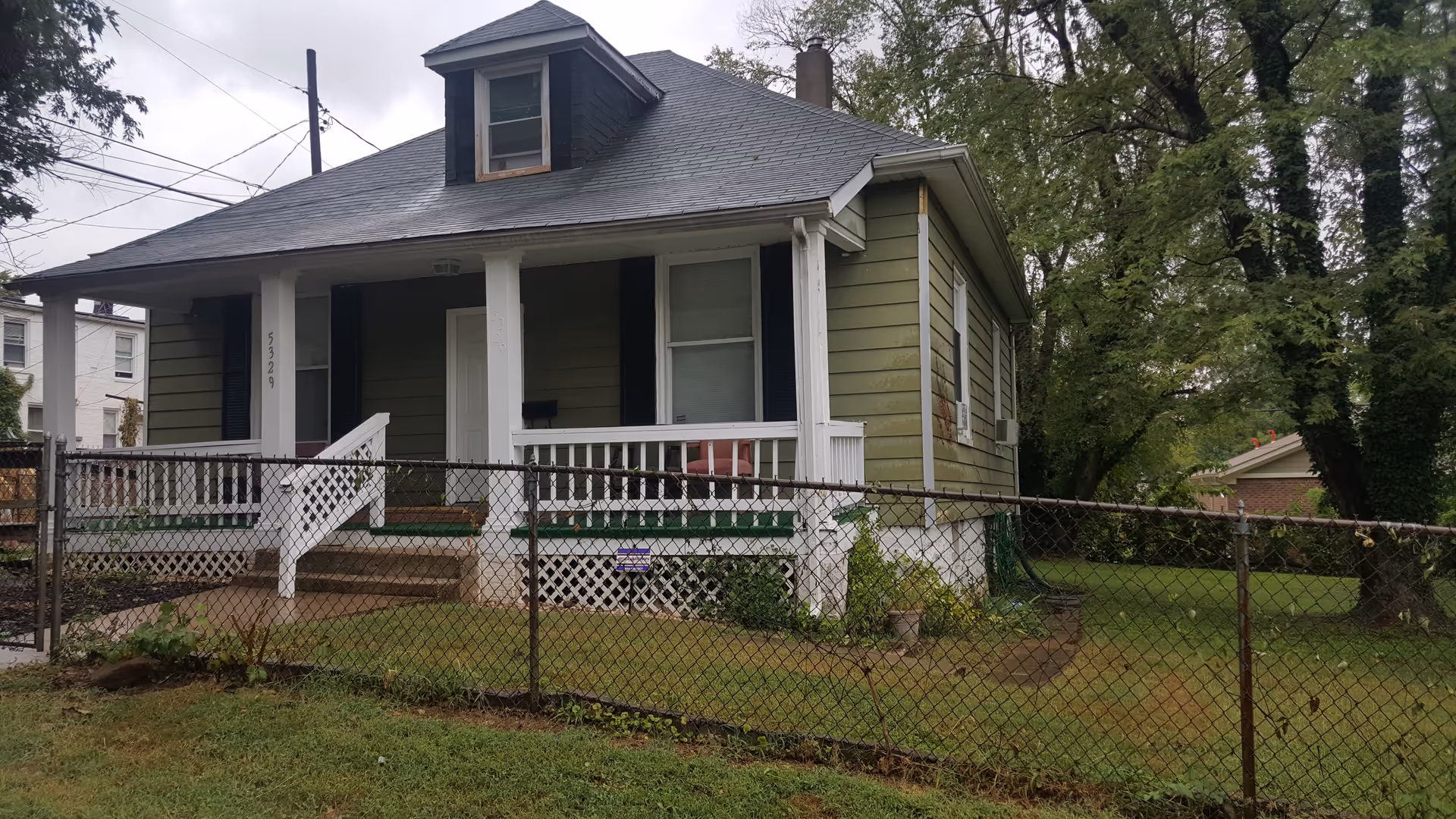A small green house with white trim and a dark gray roof, featuring a front porch with white railings and steps leading up to the door. The house is surrounded by a chain-link fence and has a grassy yard with trees in the background under a cloudy sky.