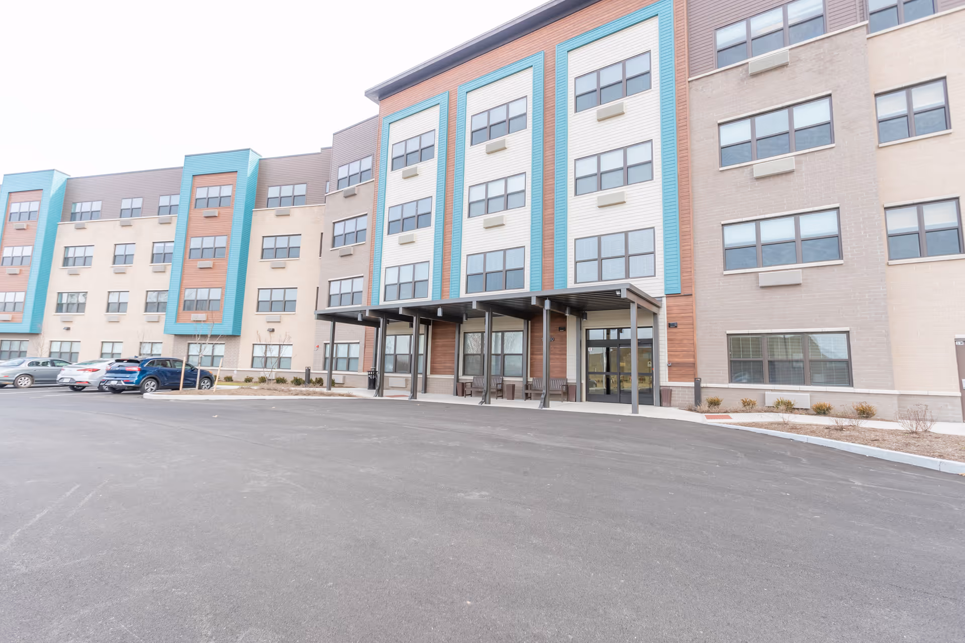 Exterior view of a modern multi-story senior living facility building with a covered entrance, several windows, and a parking area with cars in front.