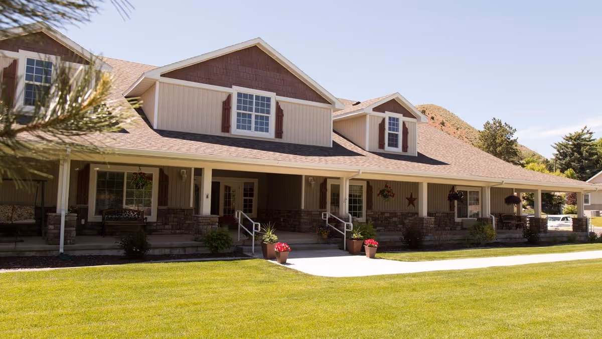 Exterior view of a single-story residential building with a covered porch, multiple windows with shutters, and a well-maintained lawn in front. There are potted plants and hanging flower baskets on the porch, with a backdrop of hills and trees under a clear sky.