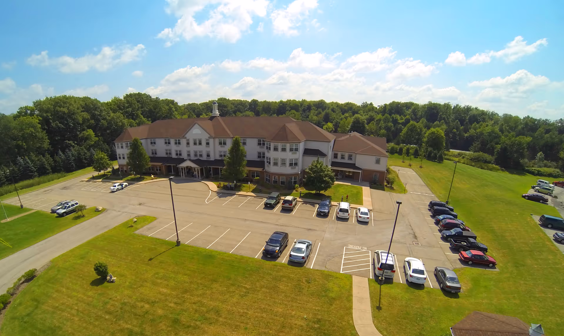 Aerial view of Liberty Arms Assisted Living facility showing a large three-story building with a brown roof surrounded by green lawns and trees. There is a parking lot with several cars parked in front of the building under a partly cloudy blue sky.