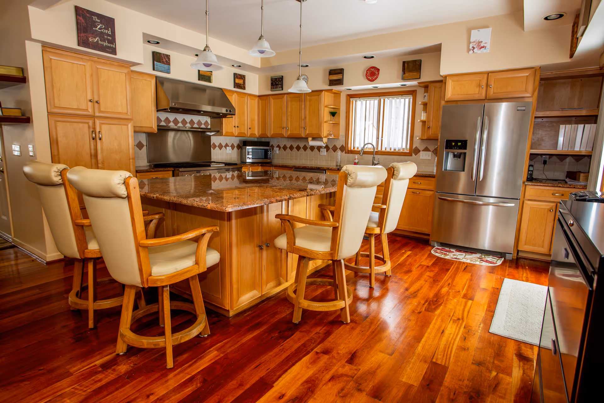 Bright kitchen with a large central granite island surrounded by four cushioned wooden bar chairs, wooden cabinets, stainless steel appliances, and polished hardwood floors.