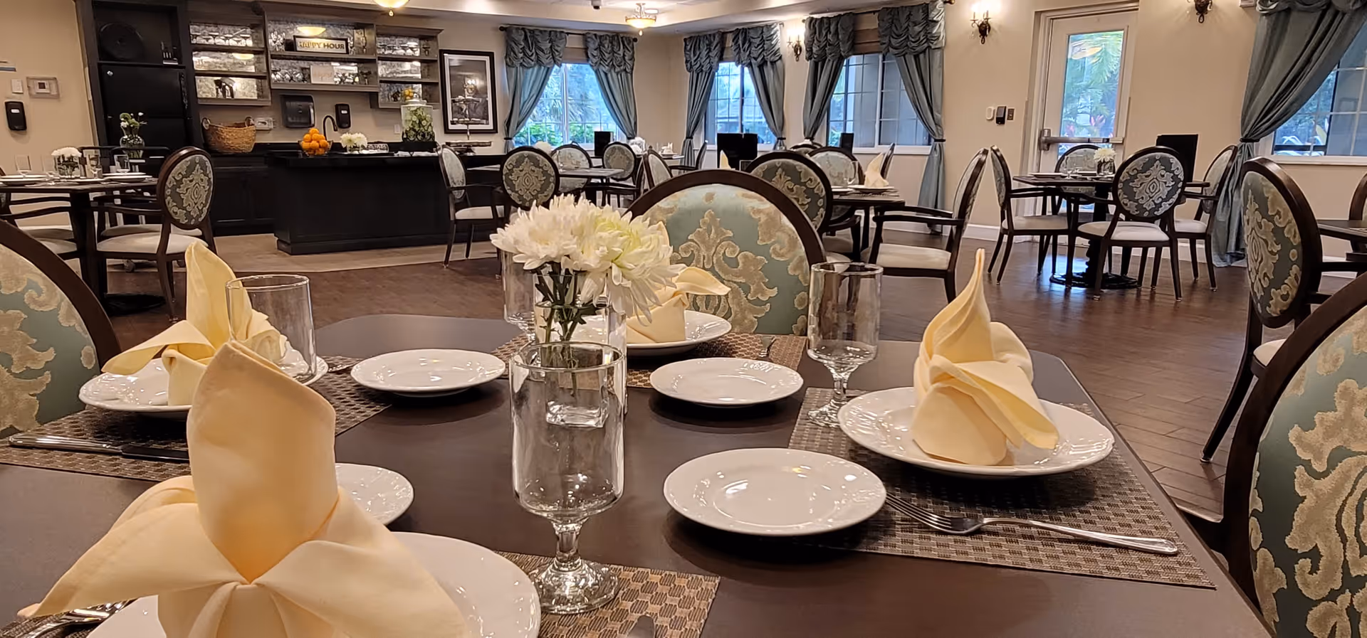 A well-lit dining room with multiple round tables set with white plates, folded yellow napkins, clear water glasses, and small vases of white flowers. The room features elegant chairs with patterned upholstery, large windows with blue curtains, and a buffet area in the background with framed pictures on the wall.
