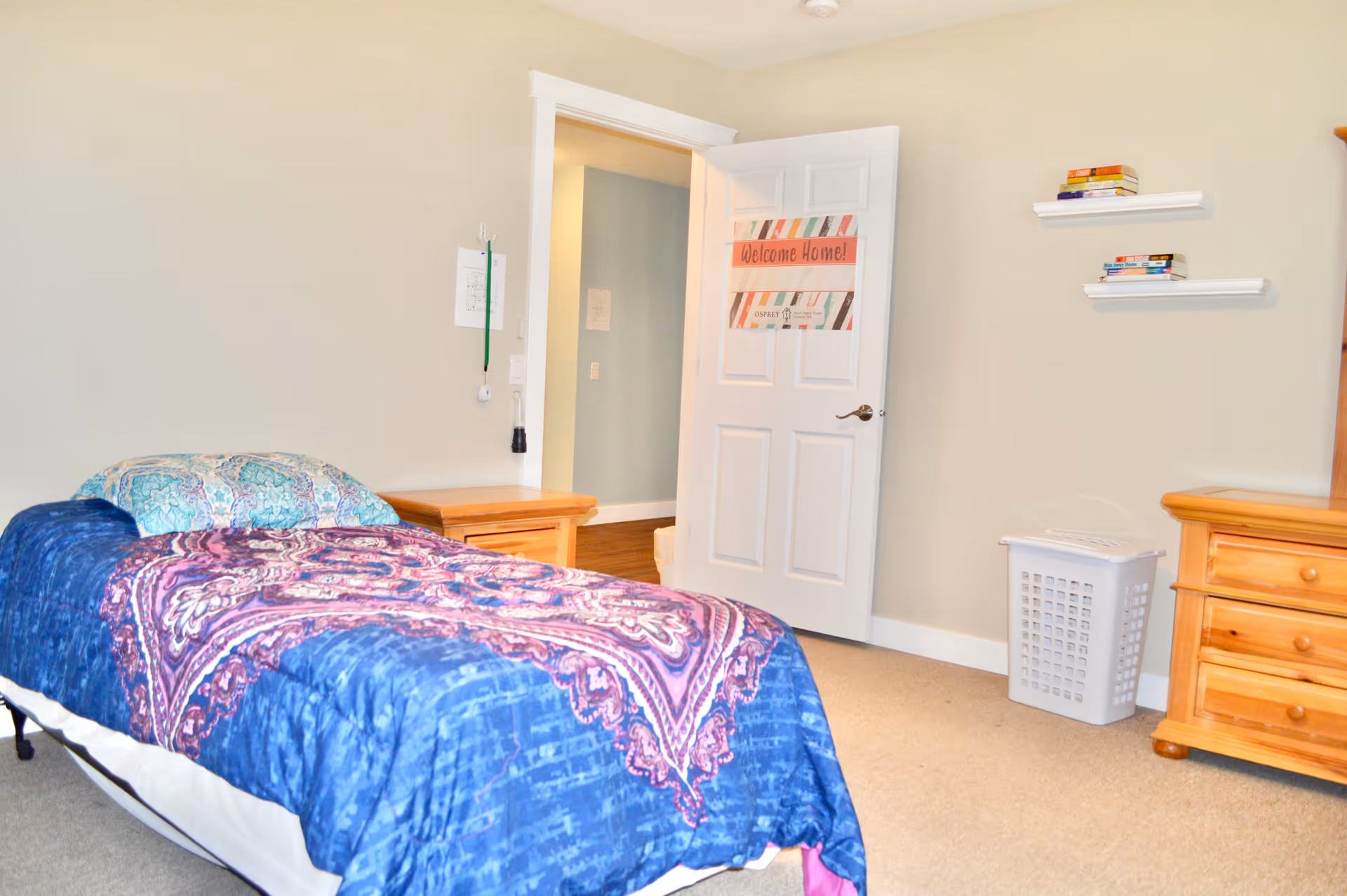 A tidy bedroom with a bed, dresser, wall shelves, and a 'Welcome Home' door.
