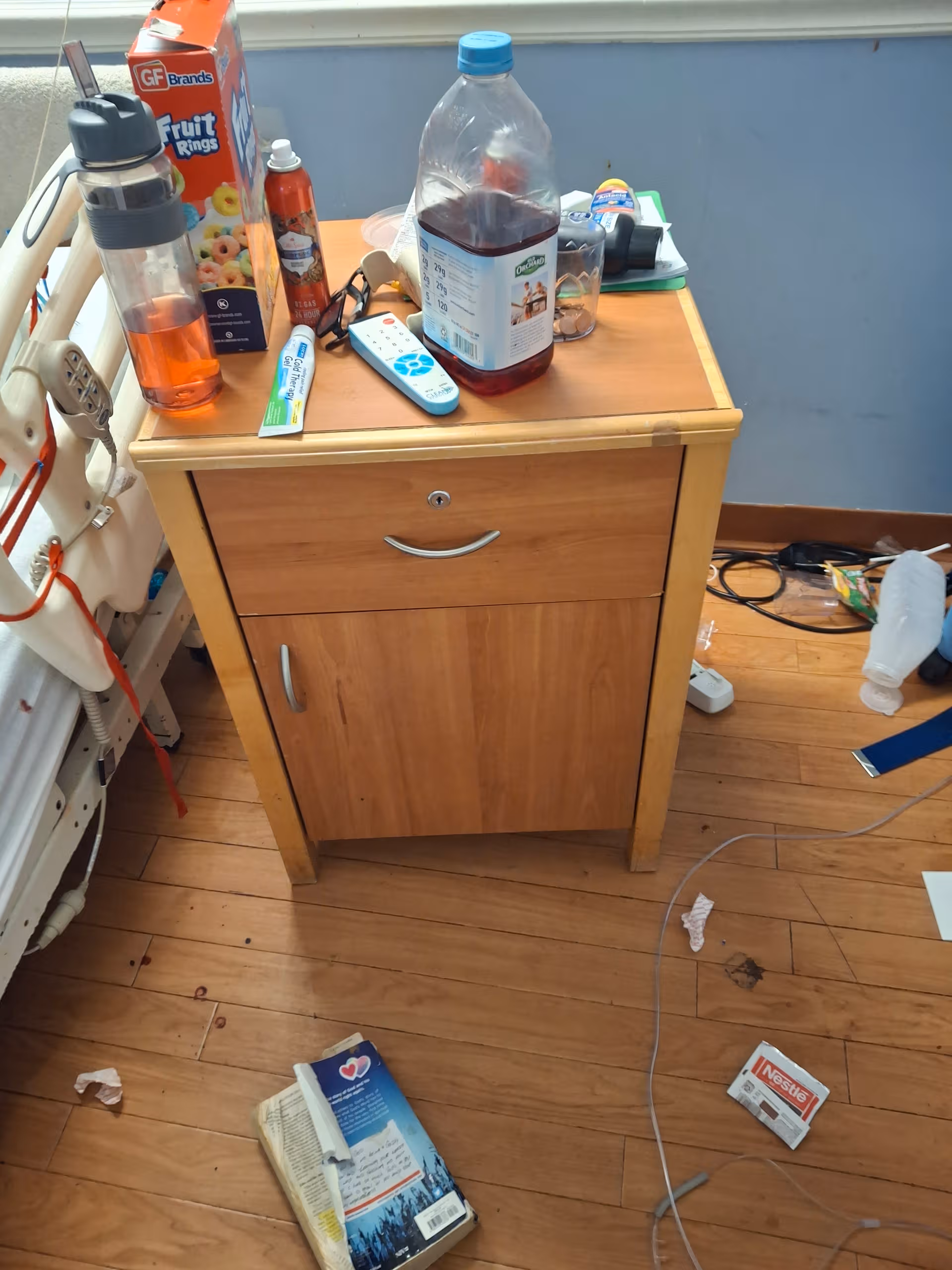 Wooden bedside cabinet in a cluttered patient room beside a hospital bed