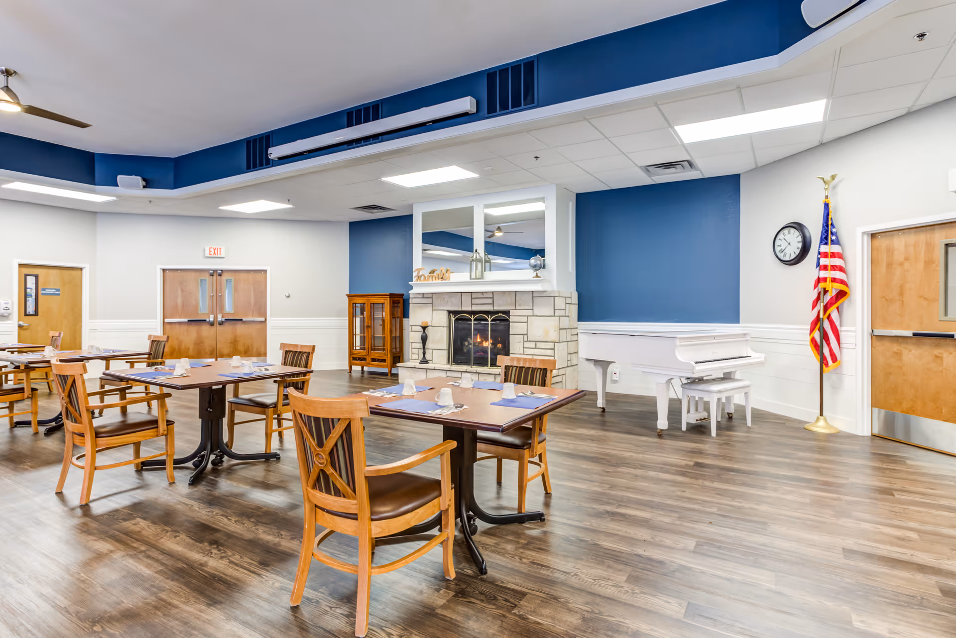 Well lit communal dining area with wooden tables, chairs, fireplace, piano, and an American flag.
