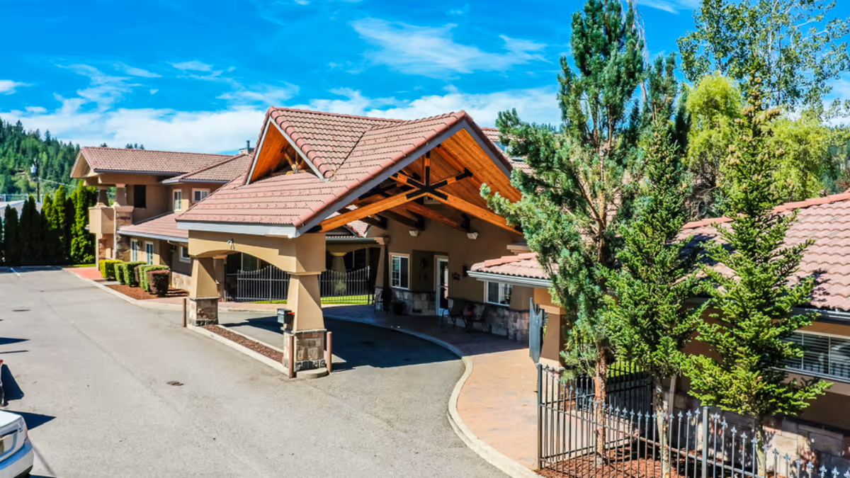 Exterior view of Pinehurst Senior Community showing a covered entrance with a tiled roof supported by stone and stucco pillars. The building has a beige facade with multiple windows and is surrounded by trees and greenery under a bright blue sky with some clouds.