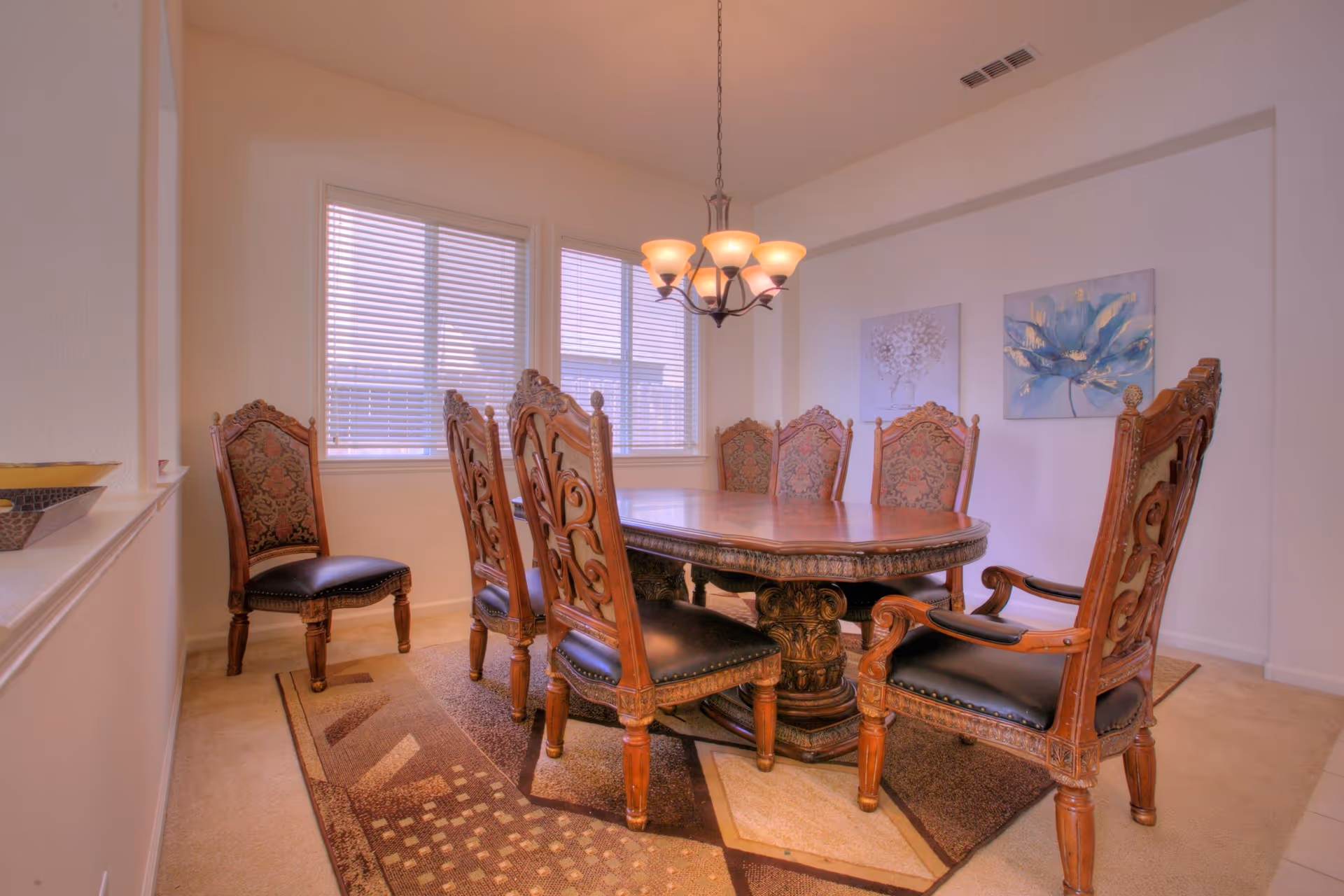 A dining room with a large wooden dining table surrounded by eight ornate wooden chairs with cushioned seats. The room has beige walls, two windows with blinds, a hanging chandelier light fixture, and two floral paintings on the wall. There is a patterned area rug under the table and chairs.