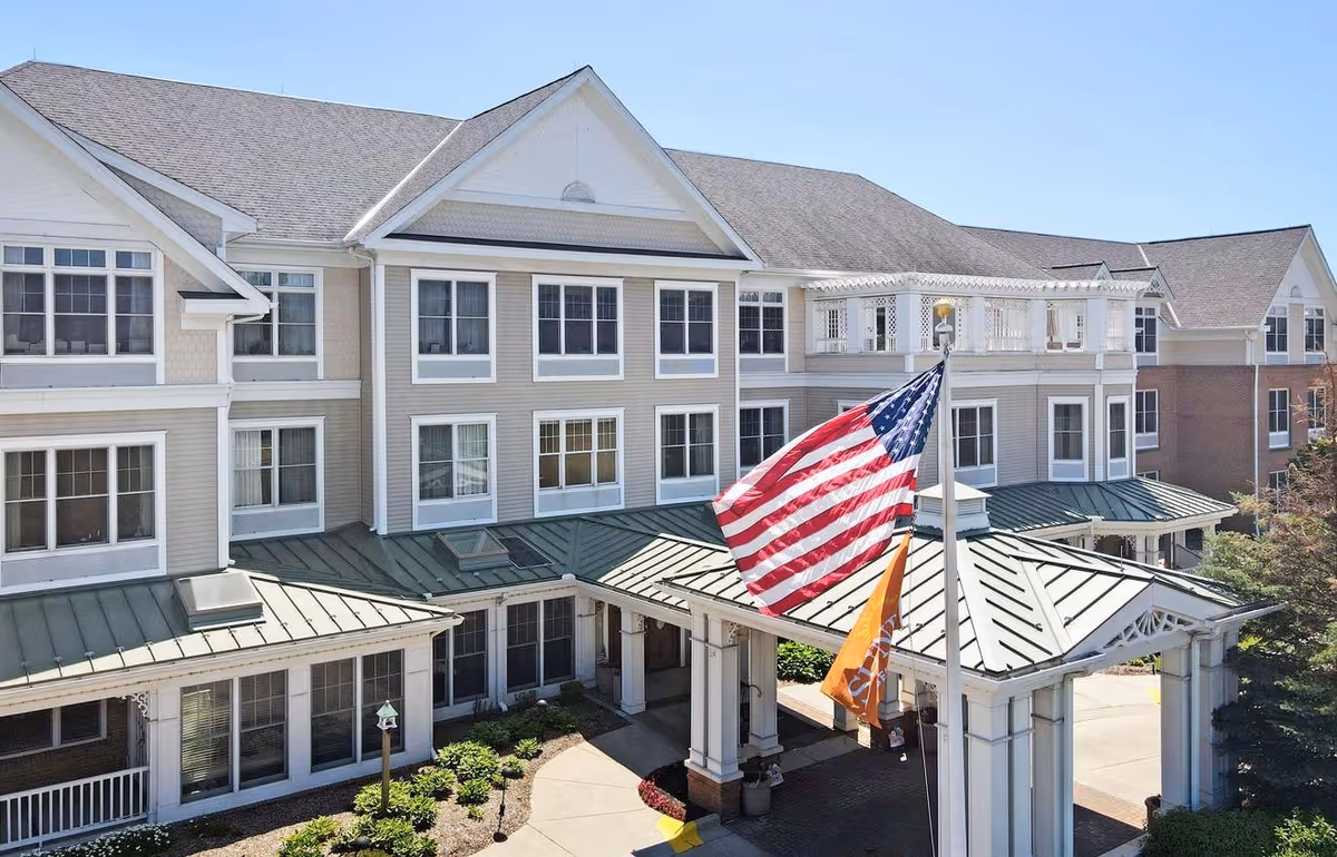 Exterior view of a multi-story senior living facility building with beige siding and white trim. The entrance features a covered driveway with a green metal roof and two flags, one American and one orange flag, flying on flagpoles. There are landscaped plants and a clear blue sky in the background.