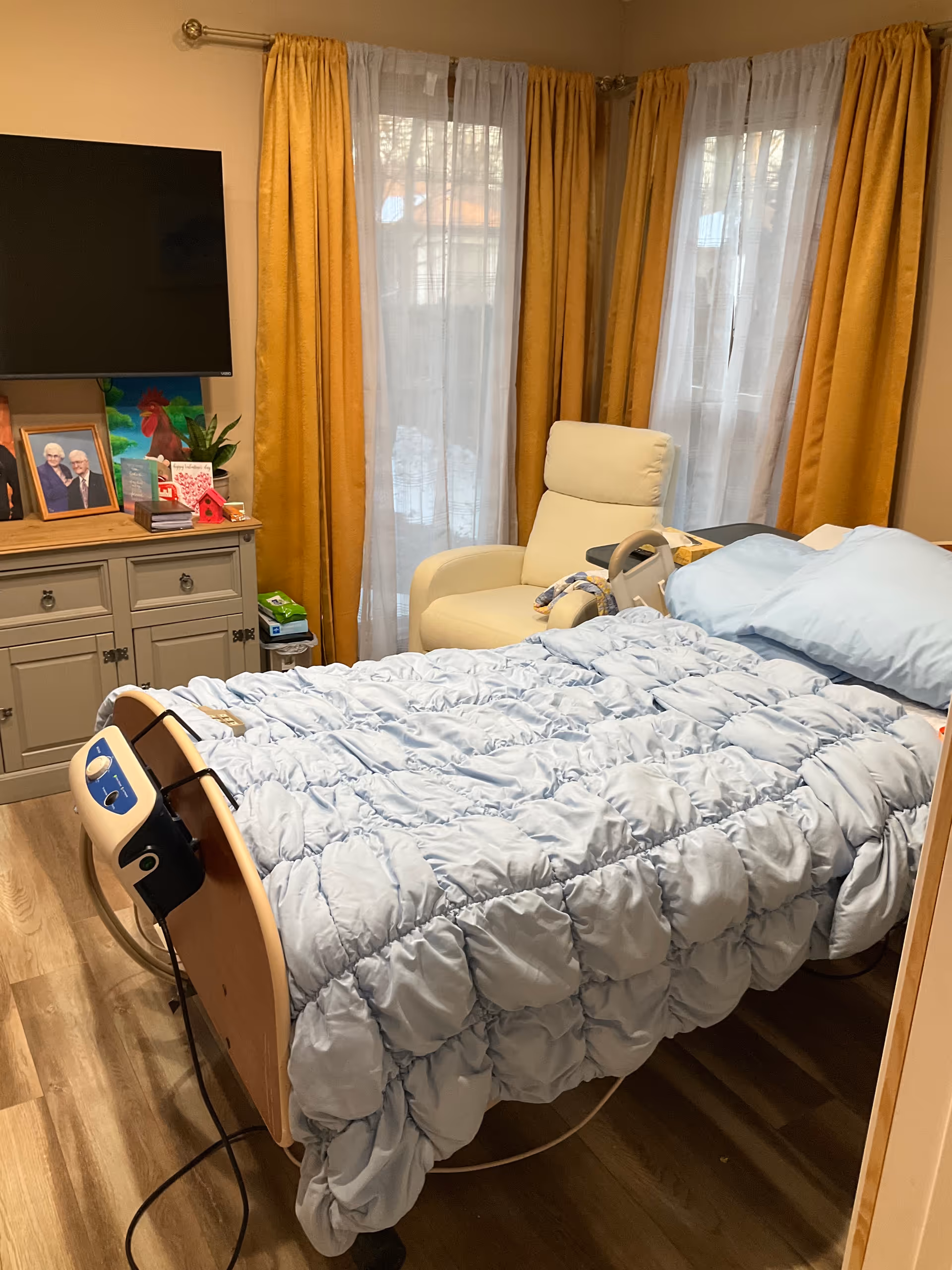 Hospital-style adjustable bed in a cozy bedroom with a TV and curtains.