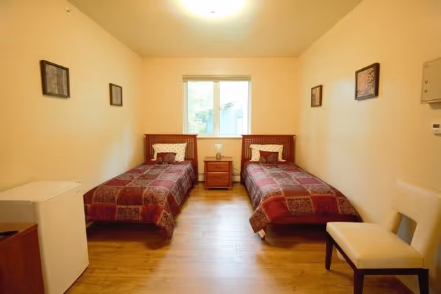 A simple bedroom with two single beds placed parallel to each other against opposite walls. Each bed has a red and purple patterned bedspread and a pillow. Between the beds is a wooden nightstand with a small lamp. There is a window centered on the back wall letting in natural light. The room has light-colored walls, wooden flooring, a white chair on the right side, and a small white refrigerator on the left side. Three framed pictures hang on the walls.