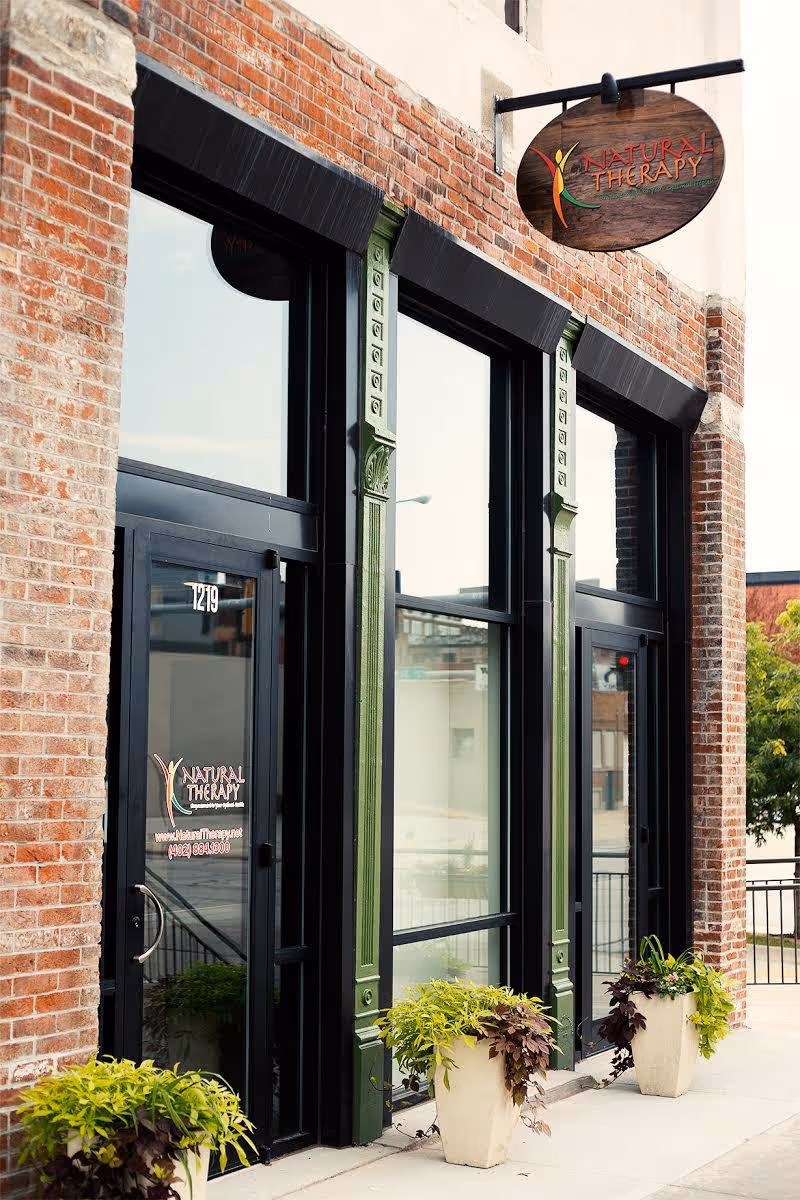 Exterior brick building entrance with green-framed glass doors and a Natural Therapy sign.