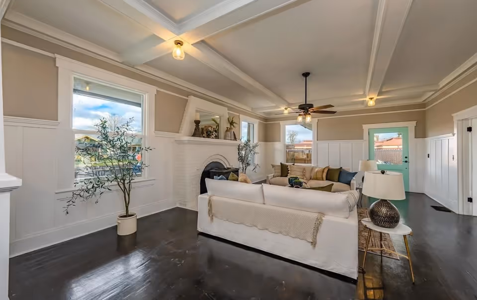 Bright living room with white furniture, fireplace, ceiling beams, and large windows.