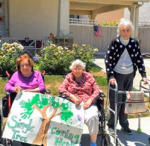 Three elderly women outside in a garden area. Two women are seated in wheelchairs holding a handmade sign with a tree and the words 'Single Tree Caring Hands'. Another woman is standing with a walker. There are flowers and an American flag in the background.
