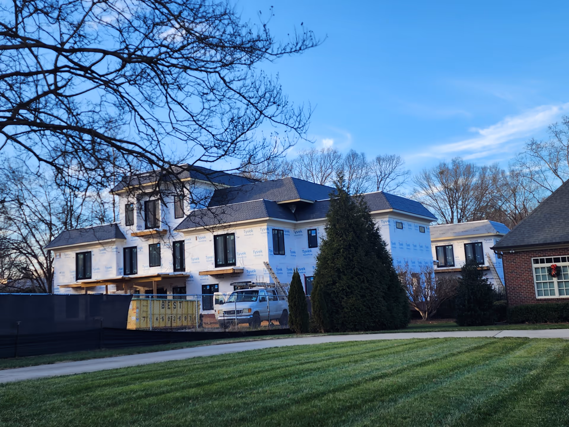 Under-construction, two-story building with Tyvek wrapping, fence, trees, and a parked truck.
