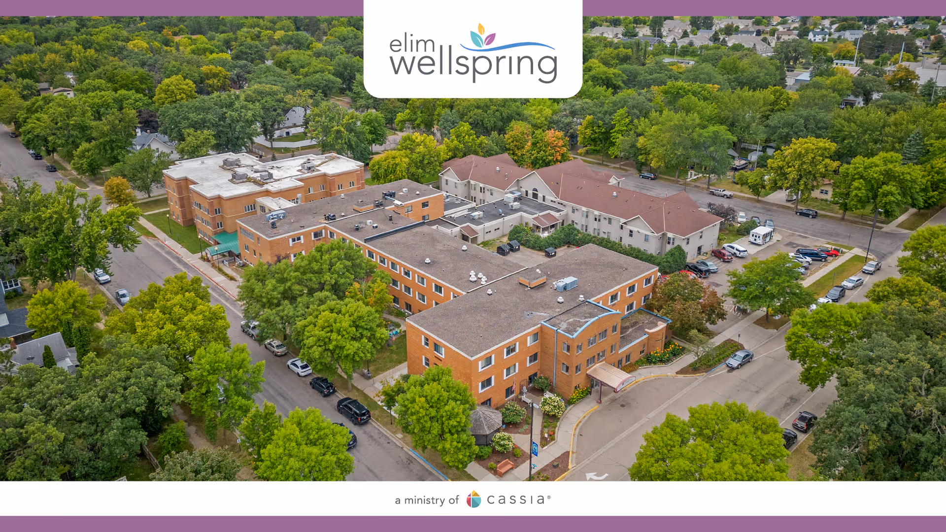 Aerial view of a brick senior living campus with interconnected buildings, trees, and parked cars.