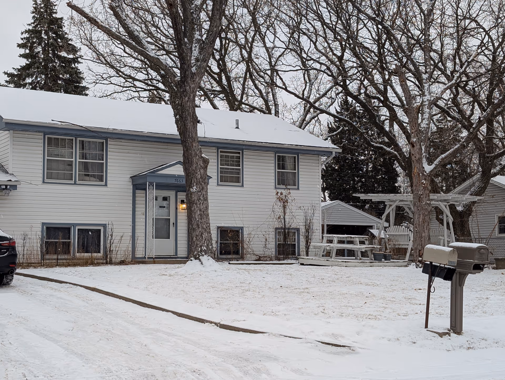 Snow-covered two-story light-gray house with a front door, mailbox, bare trees, and a picnic table in the yard.