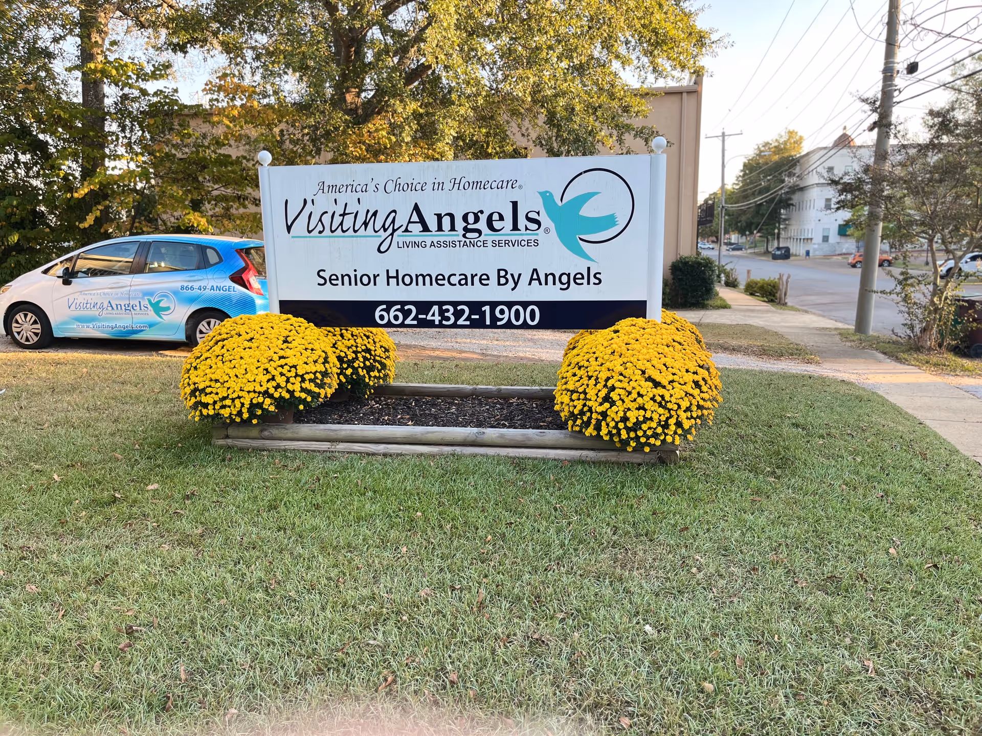 Outdoor sign reads Visiting Angels Senior Home Care Tupelo with a blue logo and chrysanthemum planters.