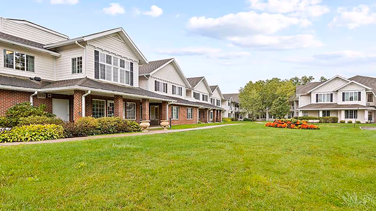 Exterior view of Holiday Fleming Point facility showing a large green lawn with a flower bed in the center, surrounded by two-story buildings with white siding and brick accents under a partly cloudy sky.
