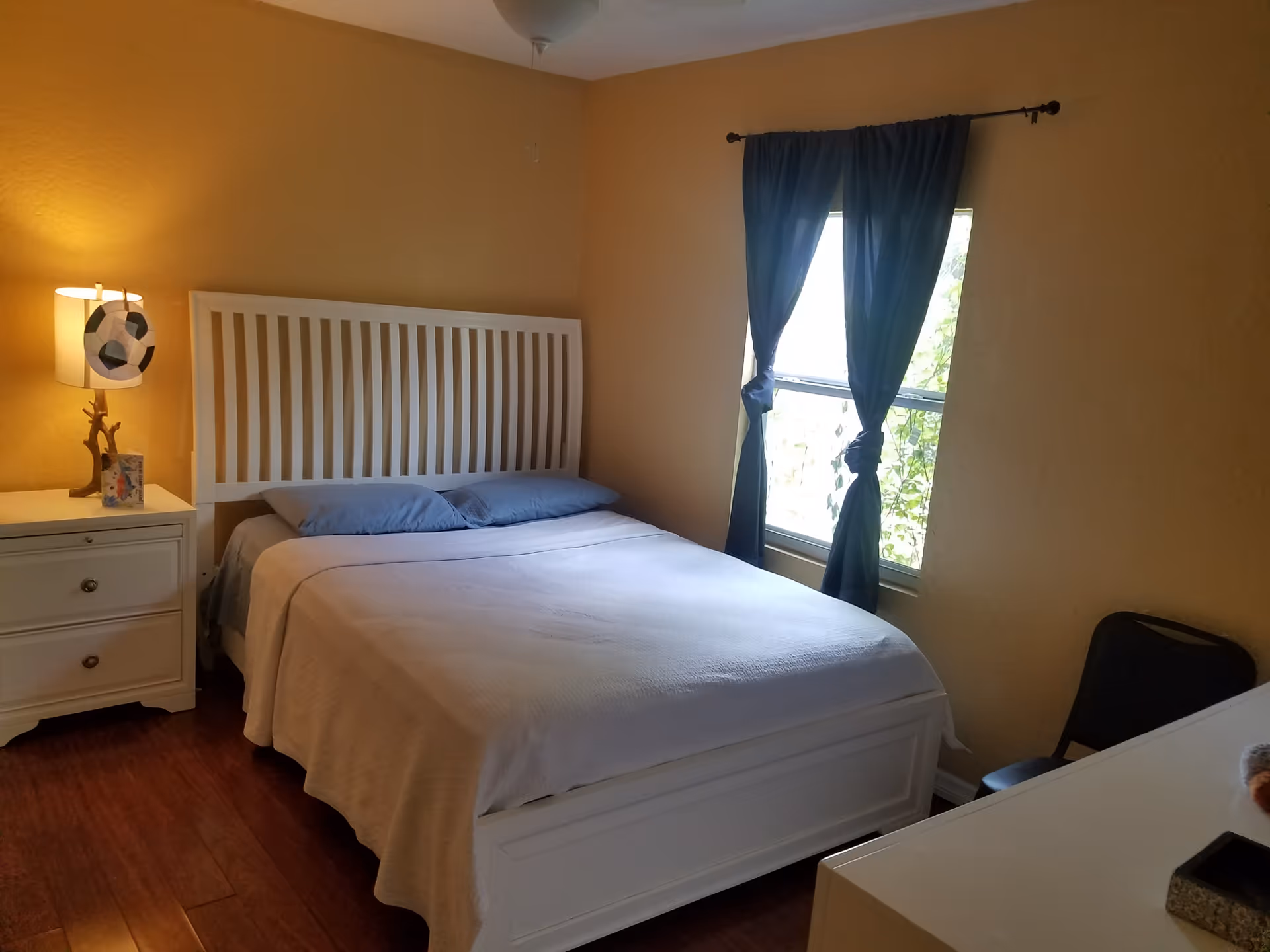 White bed with headboard, nightstand, lamp, and window with blue curtains.