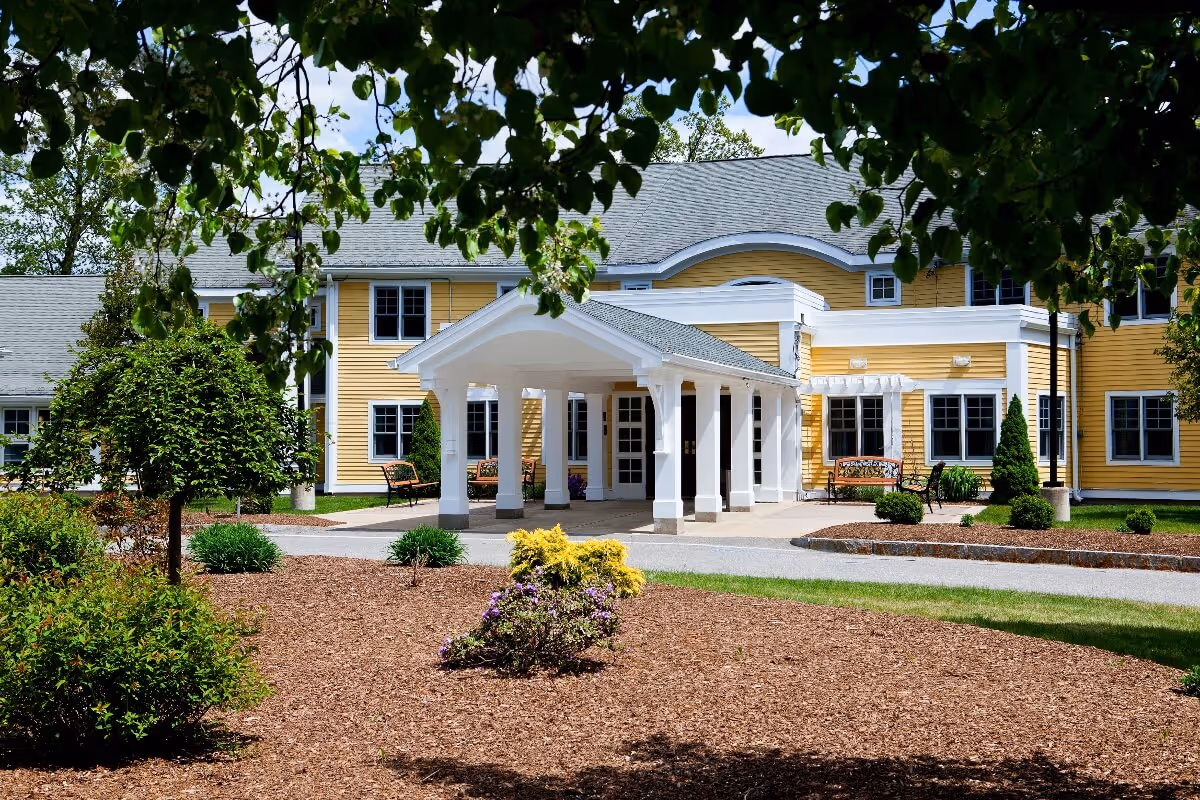 Front entrance of a yellow two-story senior living building with a white columned porte-cochère and landscaped grounds.