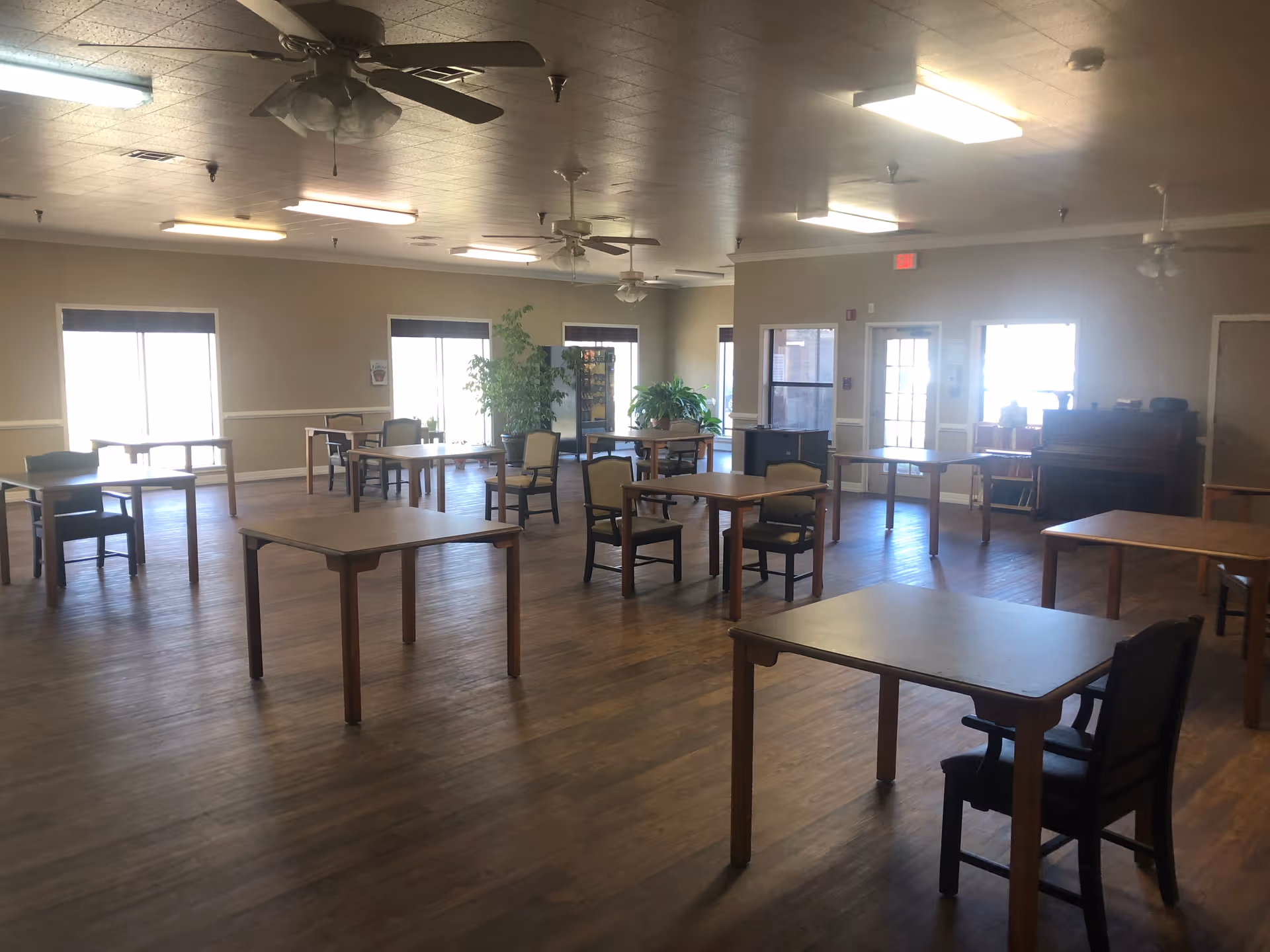 A spacious senior living common area with multiple tables, chairs, ceiling fans, and potted plants.