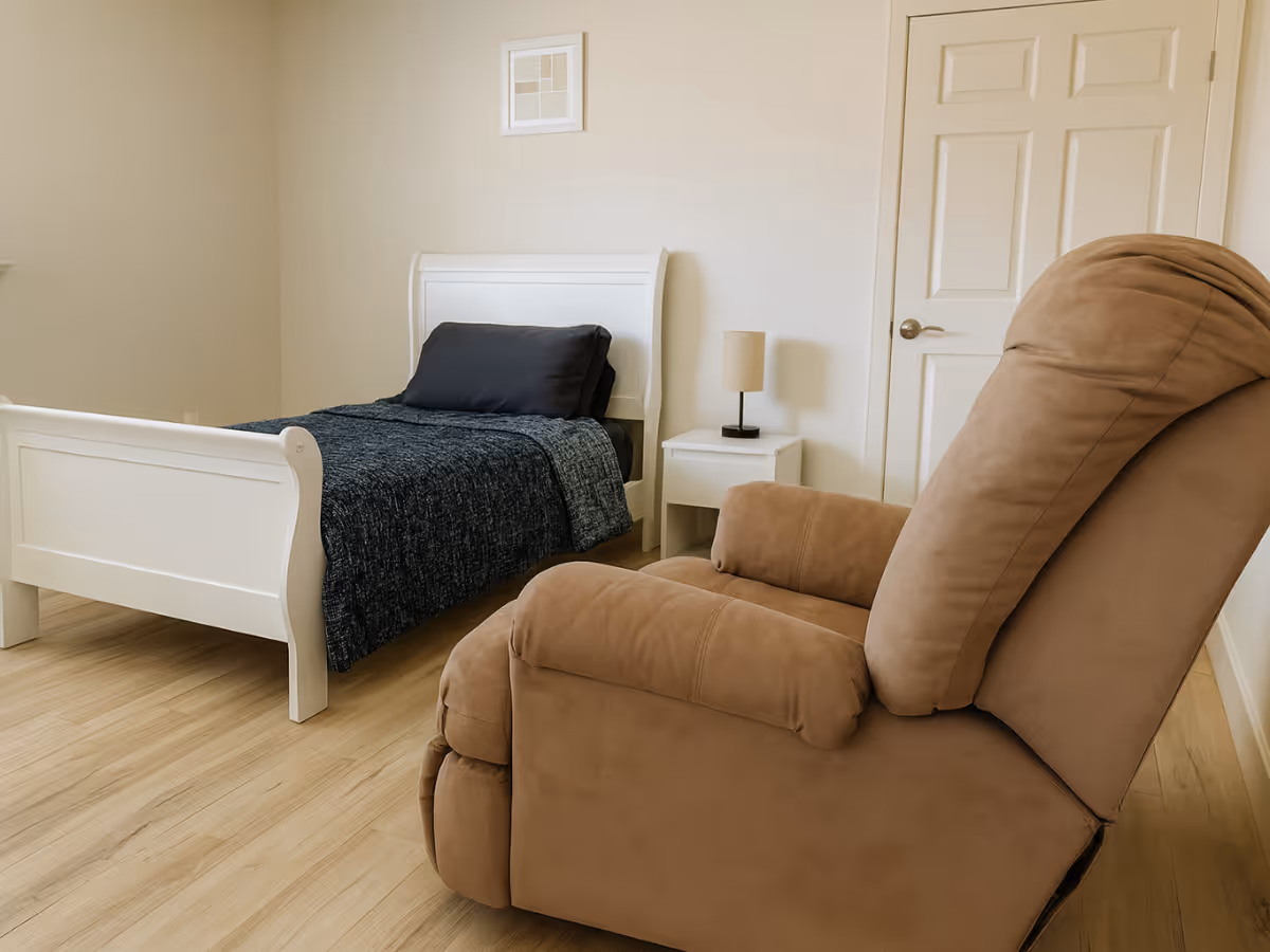 Bright bedroom in a senior facility with a white bed, nightstand, and brown recliner.