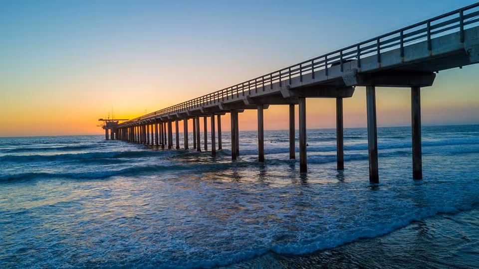 Long wooden pier extends into the ocean, ending at a glowing sunset horizon.