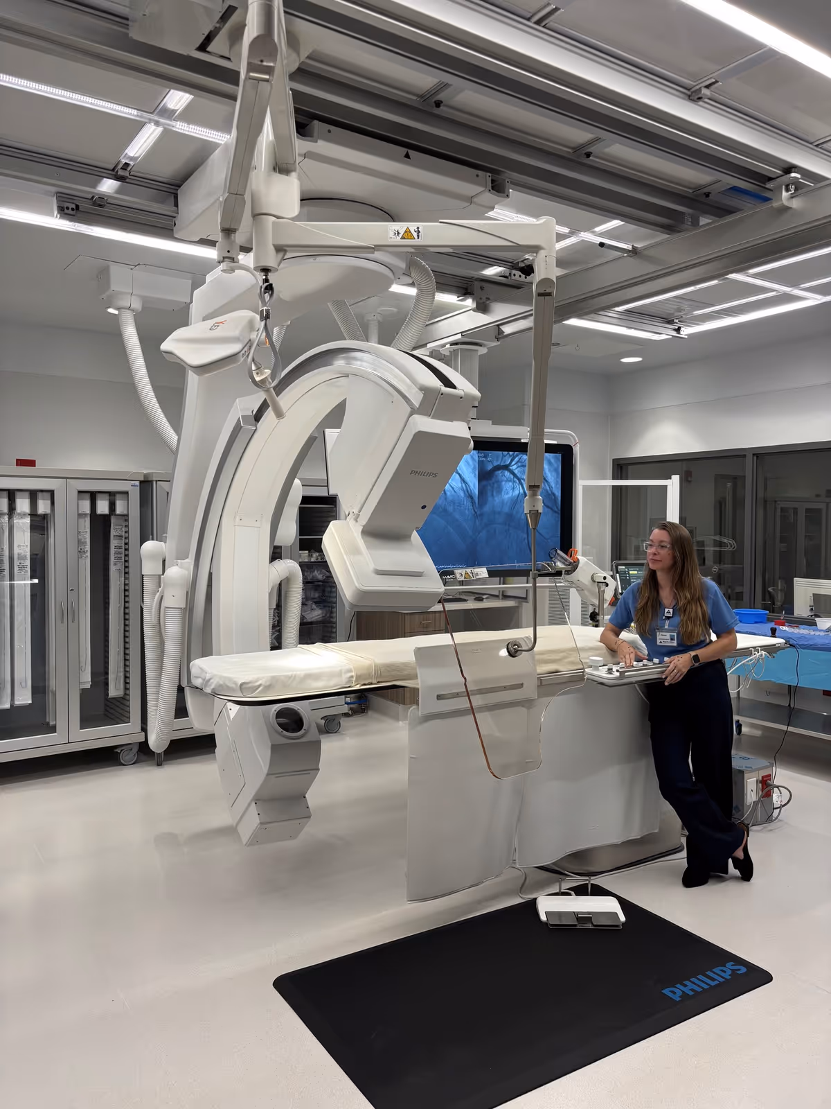 Medical imaging suite with a large C-arm machine and a clinician at a control station.