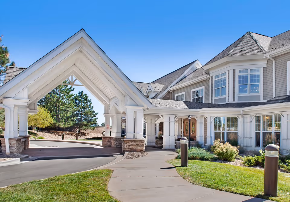 Exterior view of Hemsley House at University Park showing a covered driveway entrance with white pillars and stone bases, a curved sidewalk, well-maintained landscaping with grass and shrubs, and a large building with multiple windows under a clear blue sky.