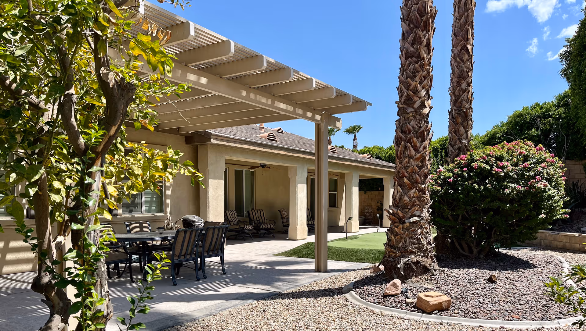 Outdoor patio area at Shiloh Senior Living in La Quinta with a covered seating area featuring a table and chairs, palm trees, a flowering bush, and a clear blue sky.
