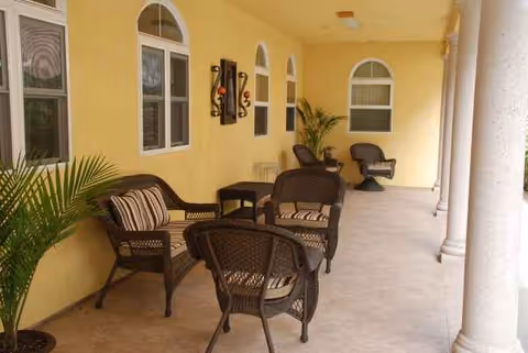 Outdoor covered patio with wicker seating, yellow walls, arches, potted plants, and columns.
