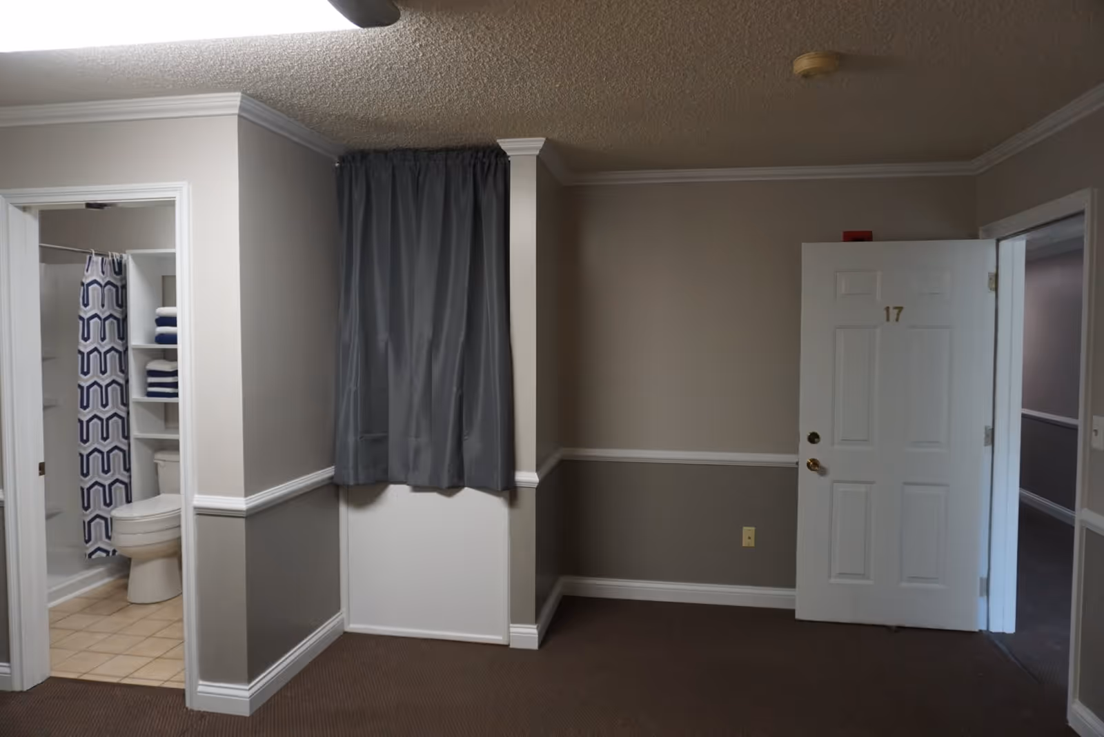 Interior view of a room showing a white entrance door numbered 17, an open bathroom with shower and toilet, and a gray curtained window.