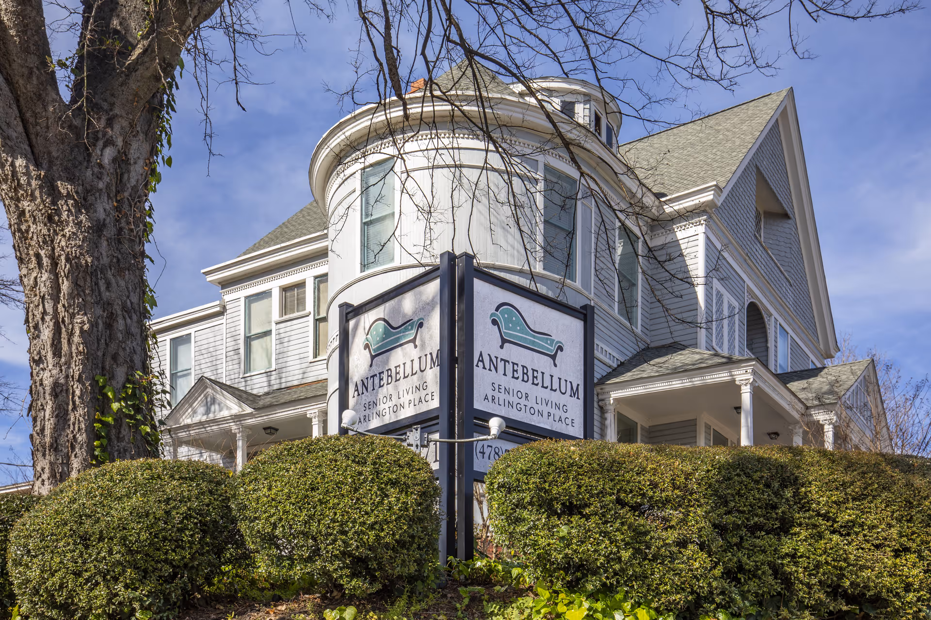 Exterior view of a large, white, multi-story senior living facility building with a rounded turret and multiple windows. In front of the building is a sign that reads 'Antebellum Senior Living Arlington Place' with a green chaise lounge logo. There are trimmed bushes and a large tree in the foreground under a clear blue sky.