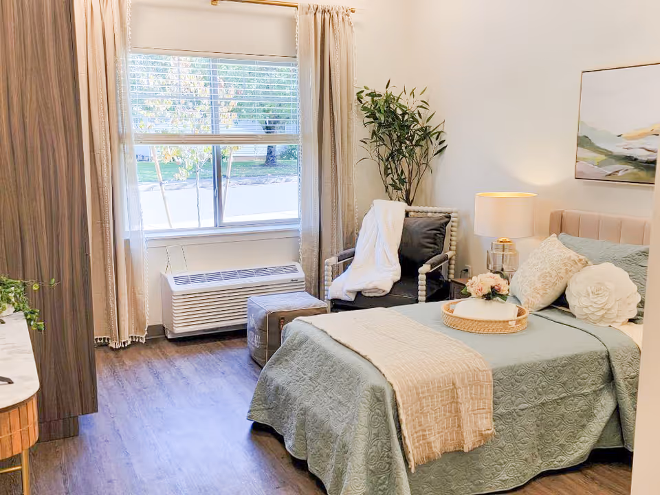 Bright bedroom with a bed, chair, plant, window, and soft decor in a senior living unit.