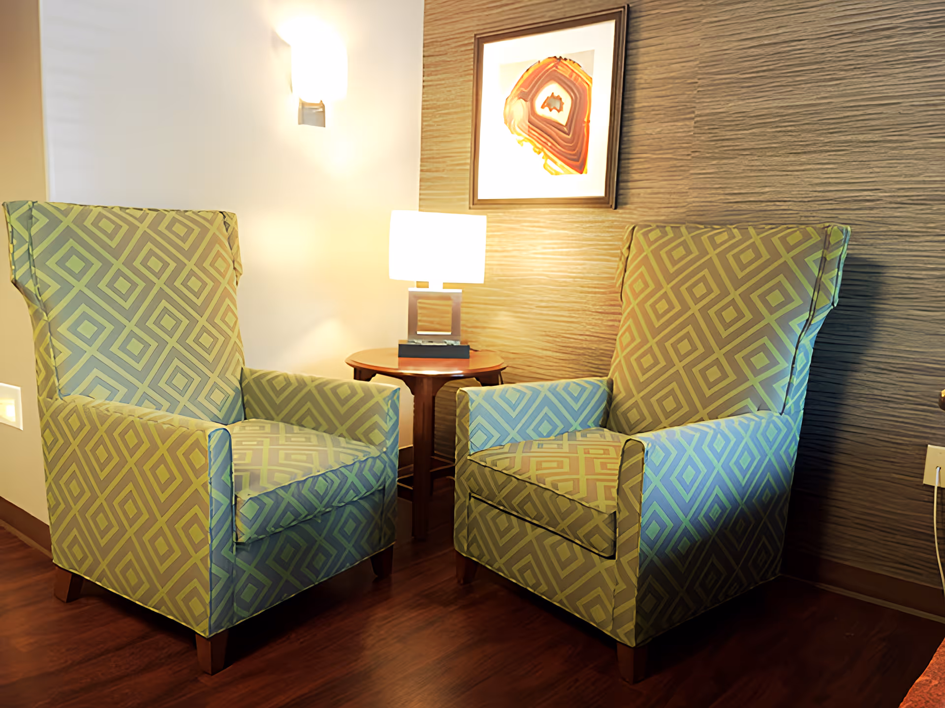 Two patterned armchairs with a geometric diamond design are positioned facing each other with a small round wooden table between them. On the table is a modern lamp with a rectangular base and a white shade. The background features a textured wall with a framed abstract artwork hanging above the table. The floor is wooden, and there is a wall-mounted light fixture on the left side.