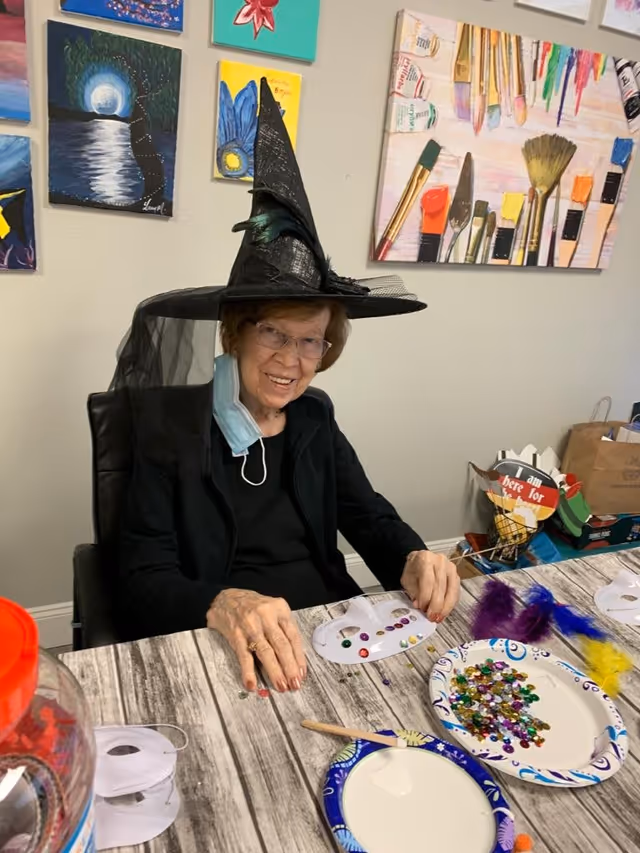 An elderly woman wearing glasses and a black witch hat with a veil is sitting at a table engaged in a craft activity. She is smiling and has a face mask hanging below her chin. The table has various craft supplies including colorful gems, feathers, and paper masks. Behind her, there are several colorful paintings on the wall, including one with paintbrushes and paint tubes.