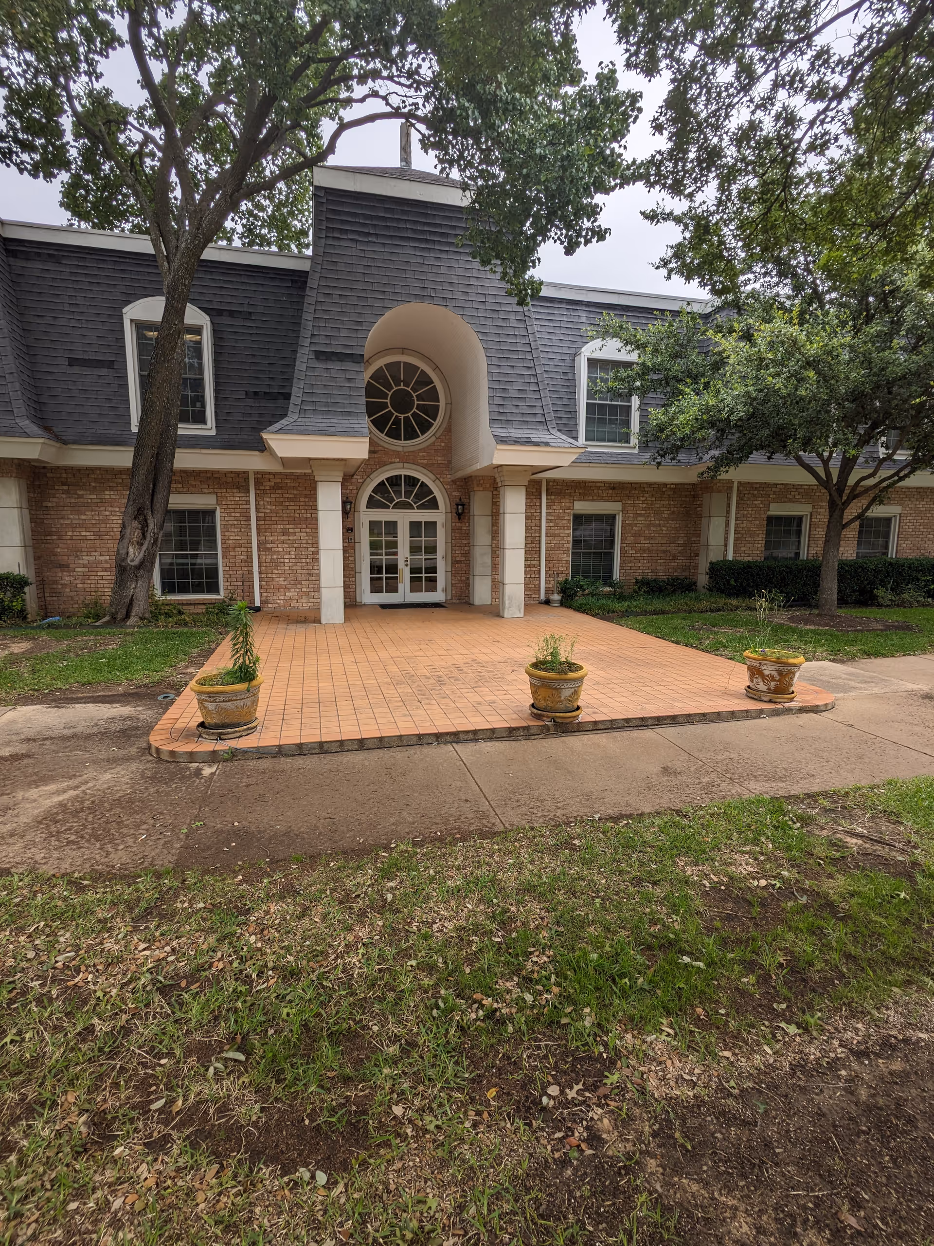 Brick building entrance with arched doorway, columns, and tiled porch with potted plants.