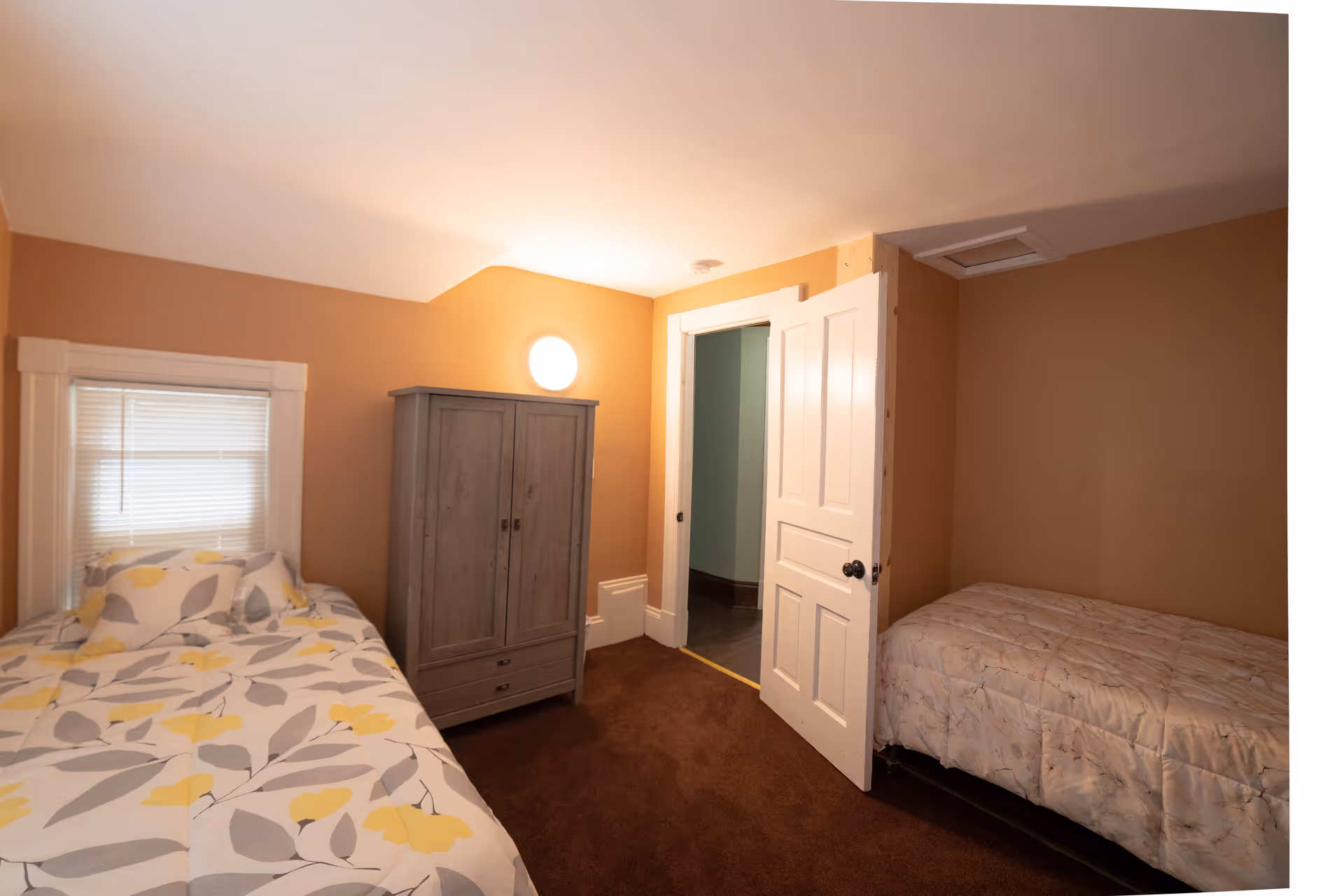 Two beds and a gray armoire fill a small brown-walled bedroom with a door.